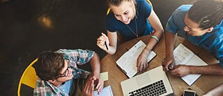 Blick von oben auf junge Leute in einem Café. Auf dem Tisch sind Laptop, Smartphone und aufgeschlagene Notizbücher.