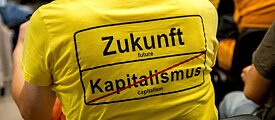 Eine an einer Tagung teilnehmende Person trägt ein T-Shirt mit der Aufschrift "Zukunft | Kapitalismus (durchgestrichen) " im Stile eines Ortsausfahrtschildes zeigt.