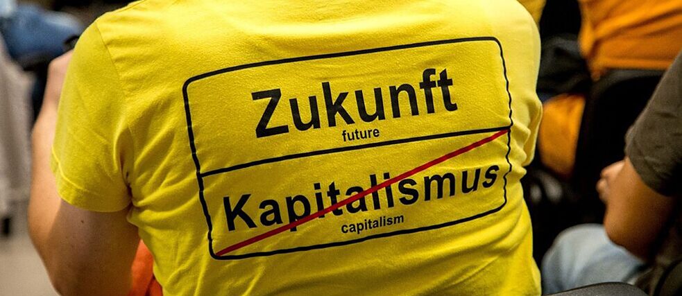 Bild von einer Zukunftstagung Eine an einer Tagung teilnehmende Person trägt ein T-Shirt mit der Aufschrift "Zukunft | Kapitalismus (durchgestrichen) " im Stile eines Ortsausfahrtschildes zeigt.