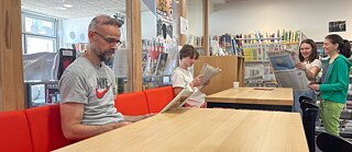 Zwei Männer und zwei Frauen lesen und unterhalten sich an großem Tisch mit Kaffee