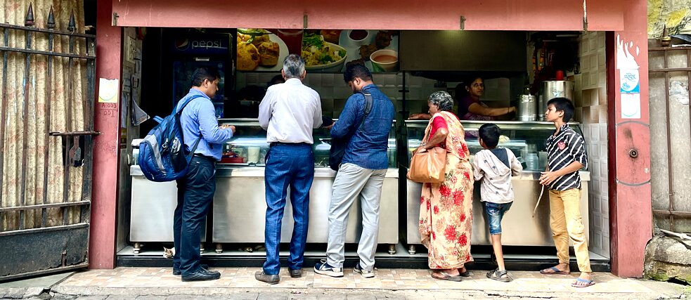 Kutumb Sakhi snack bar Six people stand in line outside the snack bar waiting for their food