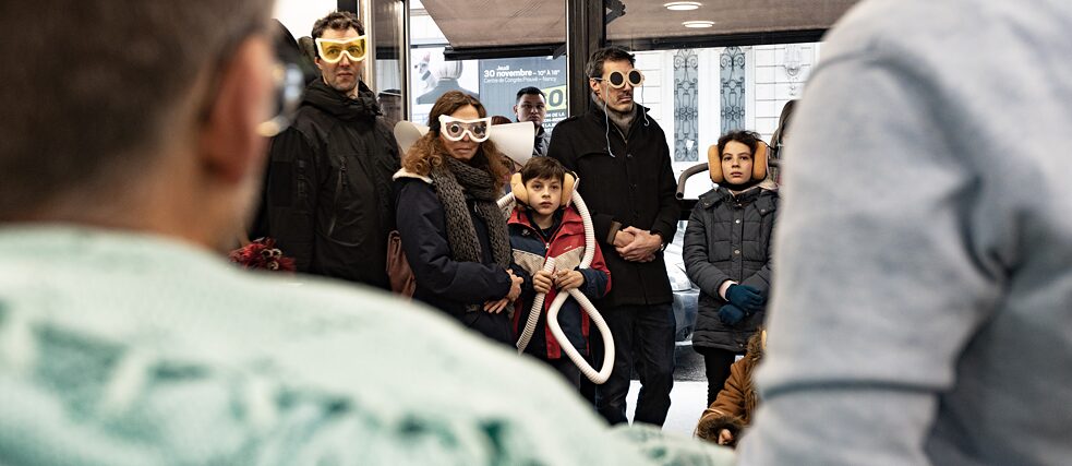 Im Vordergrund die Rücken von zwei Männern. Im Hintergrund mehrere Menschen mit skurrilen Brillen, Ohrenschützern und Schläuchen um den Hals