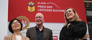 Photo : Les trois lauréat·e·s Ki-Hyang Lee, Tom Holert et Barbi Marković sont à la foire du livre de Leipzig. Ki-Hyang Lee tient un bouquet de fleurs. Sur le mur à l'arrière-plan, on peut lire ce qui suit : « 20 Jahre Preis der Leipziger Buchmesse (20 ans de prix du salon du livre de Leipzig) »