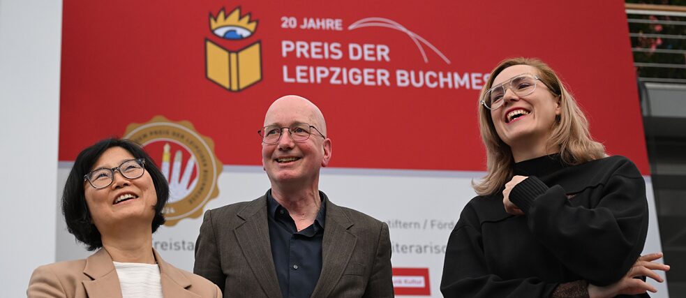 Die Preisträger*innen (l-r): Ki-Hyang Lee, Tom Holert und Barbi Marković. Foto: Die drei Preisträger*innen Ki-Hyang Lee, Tom Holert und Barbi Marković stehen lachend auf der Leipziger Buchmesse. Ki-Hyang Lee hält einen Blumenstrauß. Im Hintergrund steht auf der Wand geschrieben: "20 Jahre Preis der Leipziger Buchmesse"