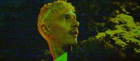 Es ist dunkel. Ein Mann mit blonden Haaren, weißem T-Schirt und blauer Jacke blickt über seine Schulter. Man sieht ihn im Profil. 