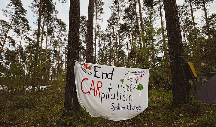 Auf dem Foto ist ein Banner, das zwischen zwei Kiefern abgespannt ist zu sehen. Darauf steht: "End CARpitalism, System Change"