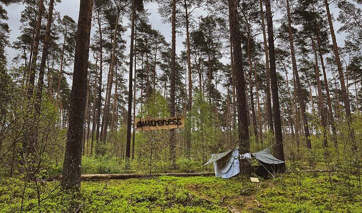 Das Foto zeigt den Awareness-Bereich einer Waldbesetzung in einem Kiefernforst. Es sind mehrere abgespannte Planen zu sehen, auf einem hölzernen Schild steht "AWARENESS".