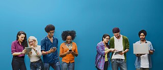 Vor einem blauen Hintergrund schauen 4 junge Menschen gemeinsam auf ihre Handys und Tablets 