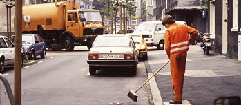 Sur l'image, on peut voir une ancienne diapositive. On y voit le service de ramassage des déchets à Dortmund. Au premier plan, une personne vêtue d'orange balaie une rue. A l'arrière-plan, un camion poubelle tourne au coin de la rue.
