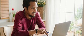 A man sits at home with his laptop and smiles at the screen.