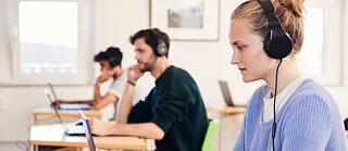 Τρεις εξεταζόμενοι με ακουστικά κάθονται σε μεμονωμένα τραπέζια και δουλεύουν συγκεντρωμένοι σε φορητούς υπολογιστές. 