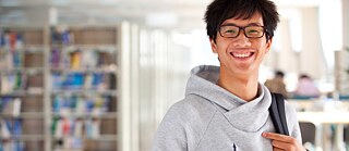 A student wearing a gray hoodie and glasses stands smiling in the library.
