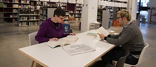 A man and a woman sit at a table in a library, both concentrating on reading.