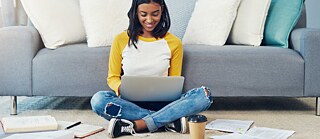 Μια νεαρή γυναίκα μελετά online στον φορητό υπολογιστή της. Κάθεται αναπαυτικά σταυροπόδι στο πάτωμα. Ακουμπάει σε έναν καναπέ. Βιβλία και φύλλα χαρτιού είναι διάσπαρτα στο πάτωμα. Μπροστά της βρίσκεται ένα χάρτινο ποτήρι με καφέ. 