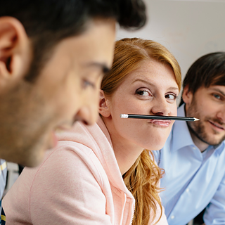 Drei junge Kollegen bei einer Besprechung. Die Kollegin macht Albernheiten mit einem Bleistift, den sie mit der Oberlippe festhält und so zum Schnurrbart macht.