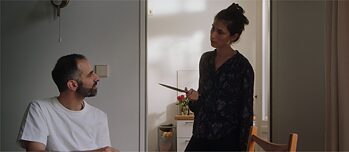 Mann sitzt und Frau kommt mit Messer aus der Küche