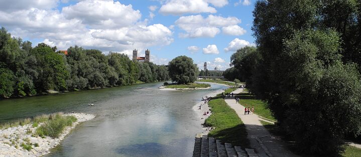 Eine Flusslandschaft mit Kiesstrand, an dem sich Menschen aufhalten. Im Bildhintergrund eine Kirche.   