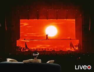 LIVEO VR-Show Im Vordergrund sitzt jemand auf einem Sofa. Die Person hat eine VR-Brille auf. Im Hintergrund sieht man die Projektion einer Show.