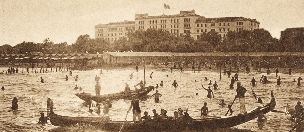 Fotopostkarte „Grandhôtel des Bains“ – Venedig, Lido Fotopostkarte „Grandhôtel des Bains“ – Venedig, Lido; Im Hintergrund ein großes Gebäude, davor ein Gewässer mit zahlreichen badenden Menschen und zwei venezianischen Gondeln