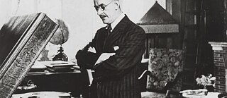 Thomas Mann, Halbprofil nach links, mit verschränkten Armen, an einem Plattenspieler stehend, Blick nach unten gerichtet.