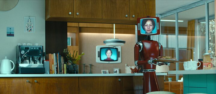 Vor dem Hintergrund einer 70er-Jahre Küchenzeile hält Cassandra - ein dunkelroter Roboter mit aufgesetzten Röhrenbildschirm, auf dem eine weiße Frau mit nackenlangen Haaren zu sehen ist - einen Teller mit belegten Brötchen. 
