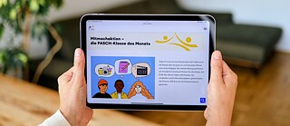 Das Bild ist aus der Sicht einer Person aufgenommen, die ein Tablet hält. Der Bildschirm des Tablets zeigt die Website www.pasch-net.de mit bunten Grafiken und Fotos.