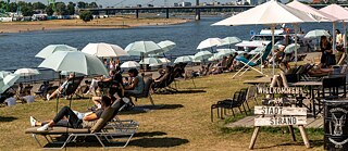 Sun loungers and parasols on the banks of a river. A sign reading ‘Welcome to the city beach’.