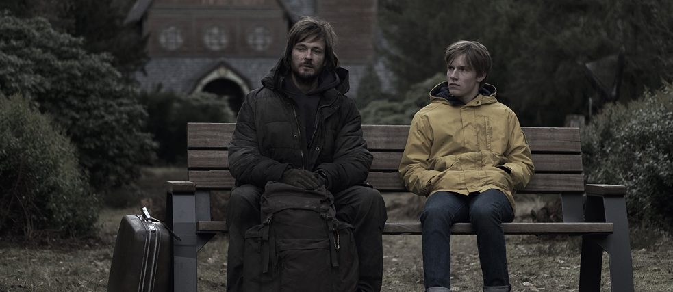 A teenage boy in a yellow raincoat and a man in dark ragged clothes sit on a bench.