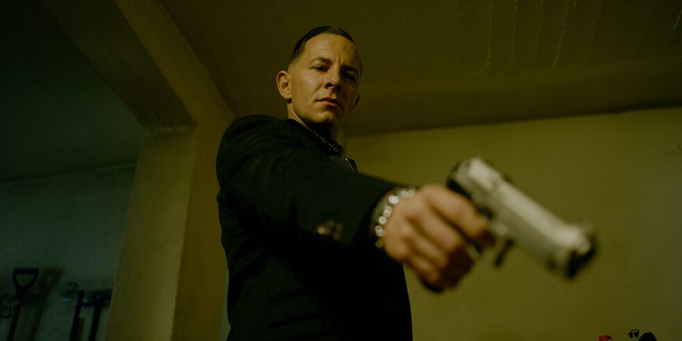 Still frame from the ZDF Series "Perfume" - "Perfume - Bondage": Bottom view of Thomas Butsche (Trystan Pütter). He stands in a room, his outstretched arm pointing his weapon diagonally down towards the camera.