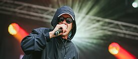 Ein Musiker mit Mikrofon, Kapuzenjacke und Sonnenbrille auf der Bühne
