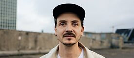 Schnurrbärtiger Mann mit Mütze auf Parkplatz neben Hochhaus