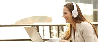 Eine Frau nimmt an einem Online Deutschkurs teil, während sie draußen an einem Tisch sitzt, einen Laptop benutzt und weiße Kopfhörer trägt.