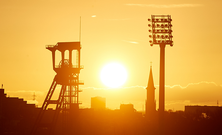 Im Vordergrund ein Förderturm der Zeche Holland. Im Hintergrund ein Flutlichtmast des Lohrheidestadions im Sonnenuntergang.