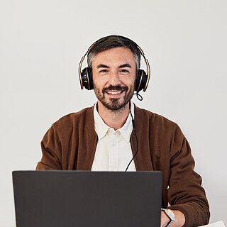 A teacher with headphones sitting in front of a laptop He is wearing a brown cardigan over a white shirt. The background is plain and bright.