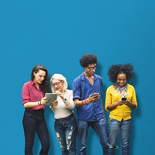 against a blue backdrop: 4 people are standing next to each other with mobile devices in their hands