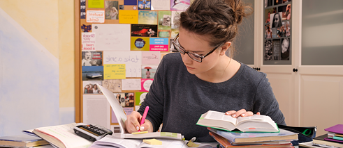 Eine junge Frau arbeitet an ihren Übungen zur Vorbereitung auf die Prüfung 'Goethe Zertifikat'. Sie sitzt an ihrem Schreibtisch und hat den Kopf über ihre Bücher gebeugt.