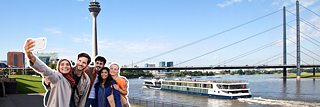 People taking a selfie on the Rhine in Düsseldorf. The Rhine Tower, a bridge and a ship can be seen in the background.